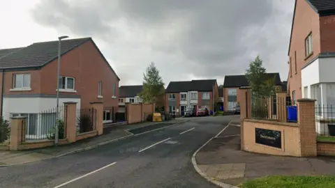 A screengrab of a Google Maps image of the entrance to the Derrymore housing development in Derry.   The estate is a mix of terraced and semi-detached homes, finished with red brick and white render. A large plaque made of shiny black stone and engraved with the placename "Derrymore" in gold lettering is recessed into a red brick wall at the entrance. 
