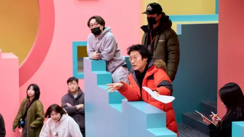 Netflix Hwang in an orange jacket on the set for Squid Game 2, adressing staff on the distinctive  staircase which is painted blue and pink  