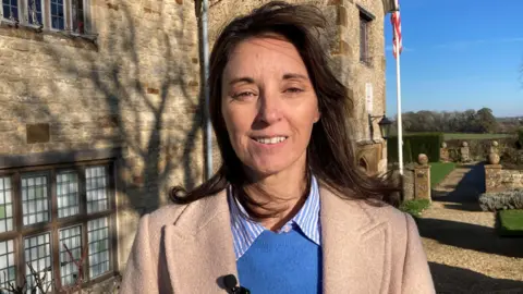 David Allard/BBC Woman in a cream coat and sky blue jumper underneath. She is smiling into the camera and has long brown hair