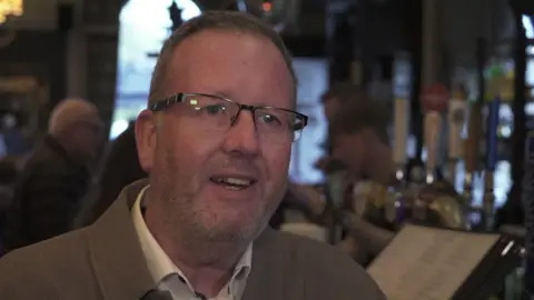 A man in glasses and a light brown jacket stands in a warm, busy bar with beer taps and people behind him.