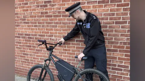 Clare Worden/BBC Ben Jarvis stands next to a black bike which has been illegally modified. The pedals are gone and a large motor is attached. Ben is wearing his black police uniform. Immediately behind him is a brick wall.