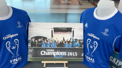 Julita Waleskiewicz Two blue Young Champions football shirts are displayed alongside a photograph of young players celebrating at the UEFA Young Champions Festival in 2013.