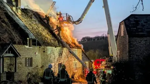 House with thatched roof - several ladders are against the roof with firefighters up ladders. Smoke and flames are spread over the roof of the home.