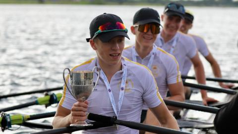 Rowers wearing white T-shirts and dark caps. All have sunglasses resting on their caps or are wearing them