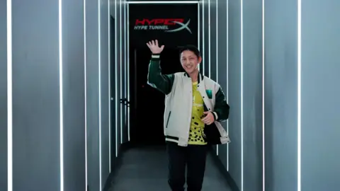 A man with short black hair emerges from the MEWC "hyperx hype tunnel," Smiling, he raised his hand to wave forward. The tunnel is brown, with white lights shining in large lines on the sides and ceiling. The man is wearing a cream varsity jacket, underneath which is a yellow eSports jersey with sponsors.