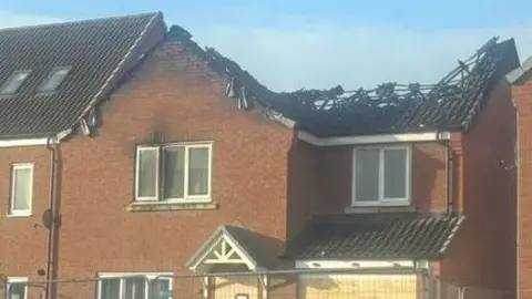 Handout A terraced house with the roof burnt down. Fencing is in front of the house. The sky is blue.