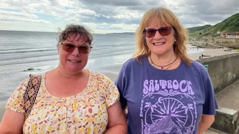 Two women stand side-by-side smiling, in front of Scarborough's South Bay beach, with the sea behind them. 