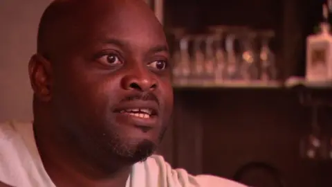Kolawole Ajay is seen a light shirt speaking indoors, with shelves of upside-down glasses and bottles blurred in the background.