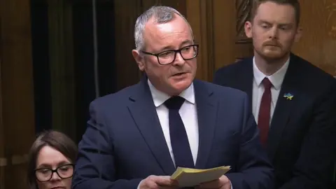 House of Commons Lee Barron in a navy suit, navy tie and white shirt speaks in the House of Commons, holding some notes. 