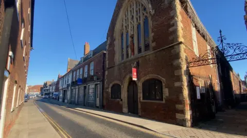 Boston Guildhall - a historical town building with dark orange brick, an arched church window and other smaller buildings next to it extending up a quiet street. The sky is blue and the sign is shining.