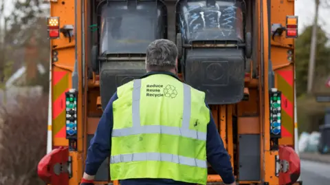 A refuse collection in a high vis vest with "reduce, reuse, recycle" on the back watching two black being being emptied into the back of an organge refuse van.