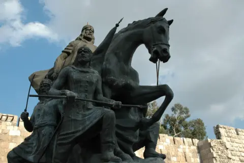 A large dark-grey statue of a man on horseback wearing medieval warrior's clothing, with two other men standing on foot beside him. The statue stands in front of stone castle walls. The sky is blue and cloudy.