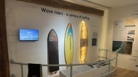 Jersey Heritage A museum with white walls. There are boards with information on. On the wall are three surf boards. In from of it are grey railings. The words " Wave Riders - A Century of Surfing" is written above the surf boards. 