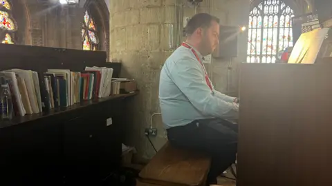 乔纳森·霍普 (Jonathan Hope) 在格洛斯特大教堂演奏电子风琴。他坐在书架旁边。