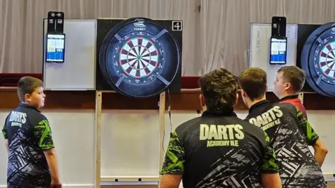 Simon Tate Four teenagers with Darts Academy North East shirts have their back to the camera and are throwing darts at a dart board. 