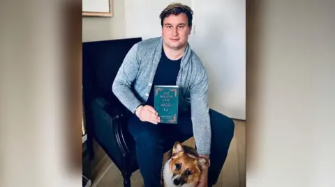 Alexander holding a copy of his book in one hand and patting his dog - a corgi - with his other hand. Alexander is wearing a blue shirt over a dark top and jeans. 