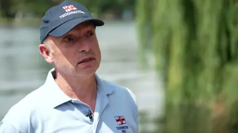 Matt Allchurch in a blue RNLI polo t-shirt and dark coloured cap. He stands in front of the river.