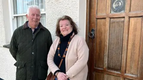 Stephen is stood on the left wearing a black quilted jacket. His hands are folded behind his back. He has white hair. Jean is stood on the right. she has her hands folded in front of her. She wears a black polo neck jumper, a cream cardigan and has a brown handbag over her shoulder. They are stood in front of a house with a brown door.
