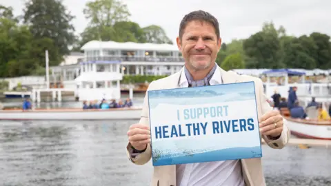 PA Media A dark-haired man in a cream blazer and white shirt stands in front of a river holding up a sign saying 'I support healthy rivers'