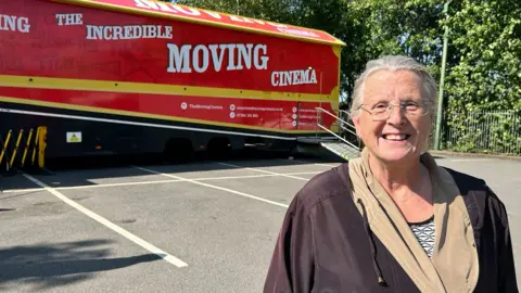 A woman with grey hair and wearing glasses and a brown and beige jacket stands in a car park in front of a red lorry with 'the incredible moving cinema' written on the side.