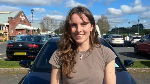 A female, wearing a grey top and necklace, smiling, standing in front of a dark coloured car, in a car park. Behind her on the right is a maroon car and on the left a black car with a blurred out number plate.