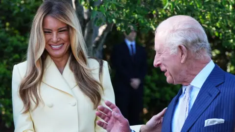 Melania Trump and King Charles laughing together at the White House. Melania Trump is wearing a cream suit and King Charles is wearing a blue suit