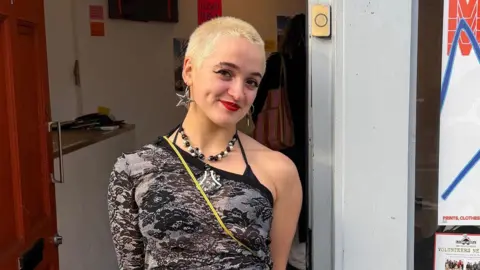 A woman with really short blonde hair, wearing an off-the-shoulder top standing outside a fashion studio