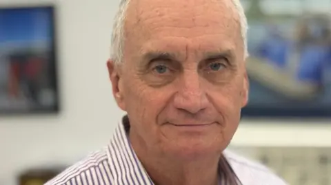 Paul Callingham is looking at the camera with a small smile. He has short white hair and is wearing a striped shirt.