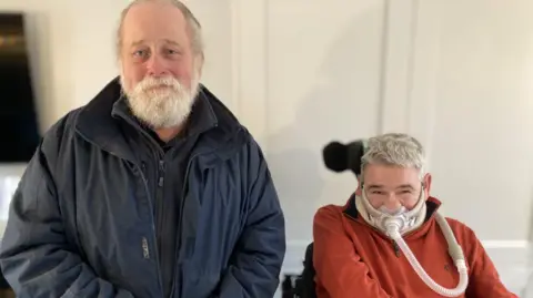 Councillor Arnold Warneken is standing on the left and is wearing a dark blue coat over a blue top. Ian Flatt is sitting in a wheelchair on the right of the image with an oxygen mask covering his mouth.