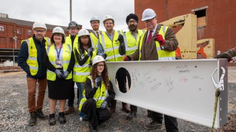 A group of people stand around a large piece of steel, they have signed their names on it. they are wearing hi-vis jackets