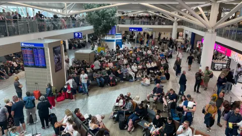 The departures lounge at Bristol Airport with people in chairs holding suitcases and watching the departures board.