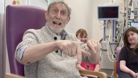 Children's author Michael Rosen, a white man with white hair and beard sat in a purple chair on a hospital ward reading to children and other young people.