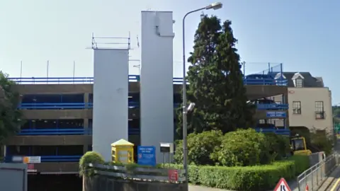 Four levels of a multi-storey car park are in the image, with blue rails along the edge of each floor. There are two floor to ceiling white towers besides the building.