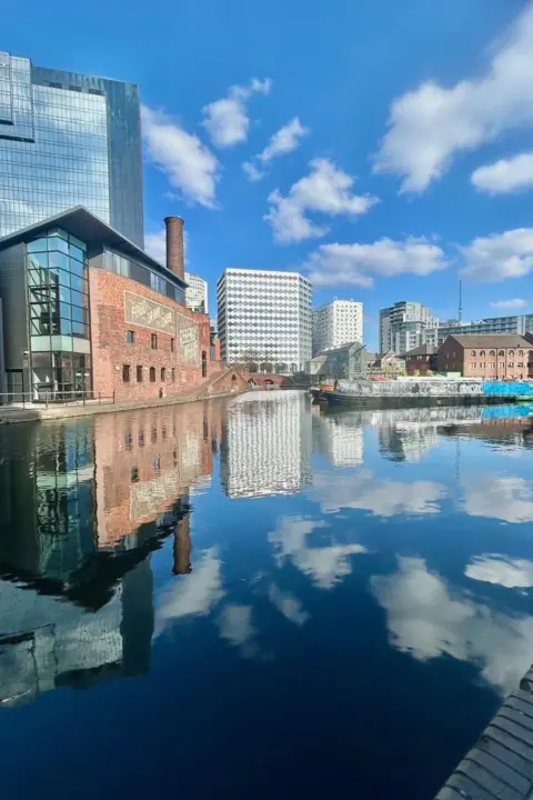 @BiteYourBrum Several buildings in the distance, beyond a canal. The sun is shining and the skies are blue with a few clouds. The buildings and sky are reflected in the water.