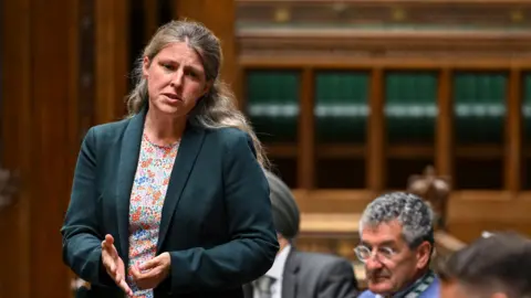 PA Media Rachael Maskell stands and gestures with her hand as she addresses the House of Commons. She has long, wavy blonde hair and wears a dark green jacket and a floral top. 