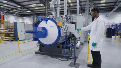 A man with black hair is wearing safety goggles along with a white lab coat and green plastic gloves. He is standing in a brightly lit industrial facility with a large cylindrical in front of him.