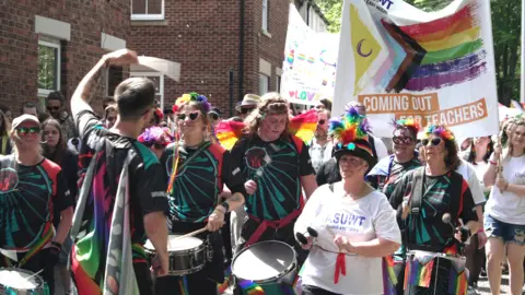 STUART PRANDLE/BBC Another shot from the march, showing drummers representing NASUWT - the teachers union. Many of them are wearing rainbow headdress - such as floral wreaths or hats. A banner has a Pride flag on it and reads: "Coming out for teachers".