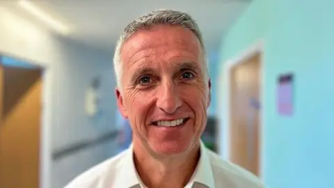A grey haired tanned man wearing a white shirt smiles.