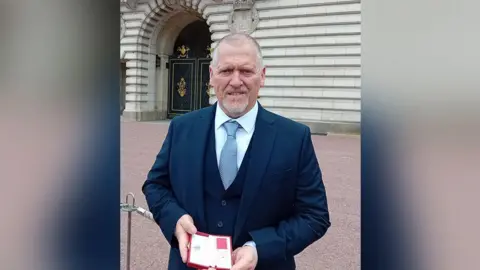 Family photograph Stephen Ornellas with short grey hair and beard wearing a smart navy suit with pale blue shirt and tie standing outside Buckingham Palace holding his British Empire Medal.