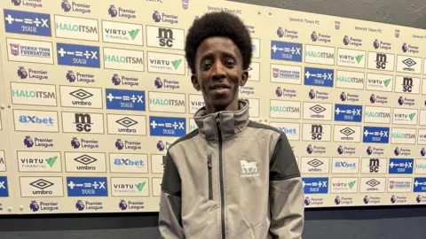 Suffolk Refugee Support A young man wearing an Ipswich Town grey and black hooded top. He is standing in front of an advertising board featuring a variety of different logos.