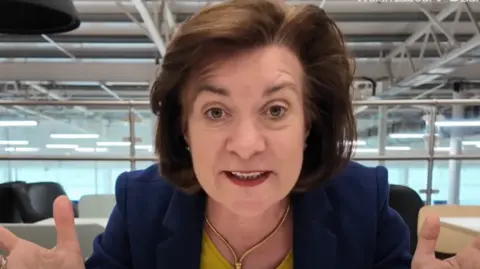 Eluned Morgan, a middle aged woman with brown hair, gestures as she speaks to the camera.