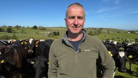 Robert Craig, a man with balding, short hair, wearing a green fleece and navy t-shirt, looks at the camera. A herd of black and brown cows are behind him in rolling green fields. It is a sunny day with blue skies. 
