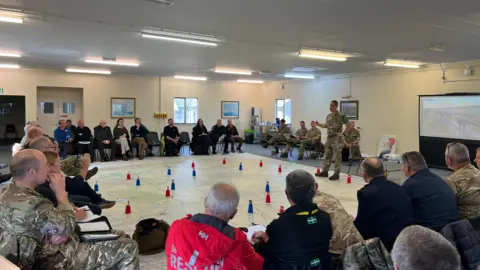 Col Jim Bird stood in front of a group of people delivering a presentation about the plans for the challenge. He is stood on a large map with different pointers dotted around it. There is also a small screen behind him with an image.