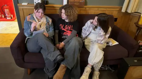 Ross Pollard/BBC Three young women sit on a dark brown sofa inside a recording studio. One of them is writing on a notepad and the other two are looking at her. All three are dressed in casual clothing