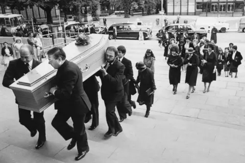 Getty Images Pall bearers carry a coffin at the funeral of Dr Amal Dharry up the steps of Coventry Cathedral, with mourners following on behind with funeral cars parked in the background