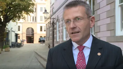 BBC Philip Ozouf stood to the right wearing a black suit jacket, a white shirt and a red and white spotted tie. In the background is a light brown building with an arched doorway and a pink and grey brick building behind him. 