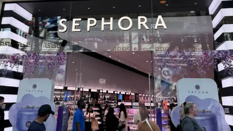 The front of a Sephora shop, it has large glass windows and doors with a sign above that says Sephora. Inside people can be seen shopping