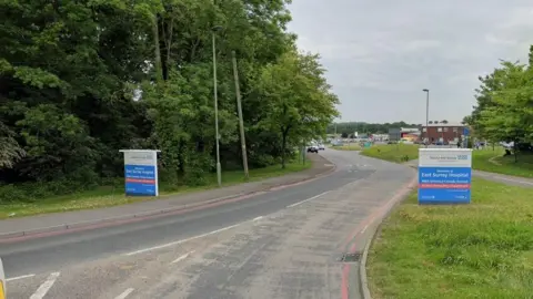 Google The road leading in to East Surrey Hospital with blue signs and the name of the hospital on the side of the road