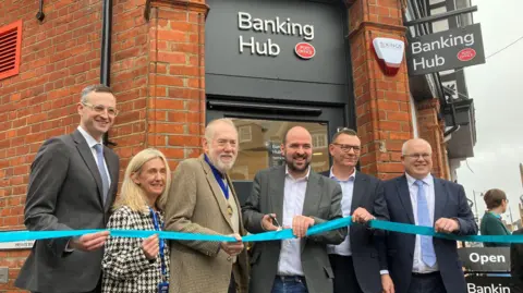 Joshua Holmes-Bright/BBC Five men and a blonde woman stand in a line holding a sky-blue coloured ribbon. They are smiling. Behind them is a bricked building, and the sign above reads BANKING HUB. It is a cloudy day.