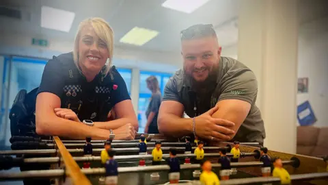 A policewoman with blonde hair leans on a football table next to a man. They both smile at the camera.
